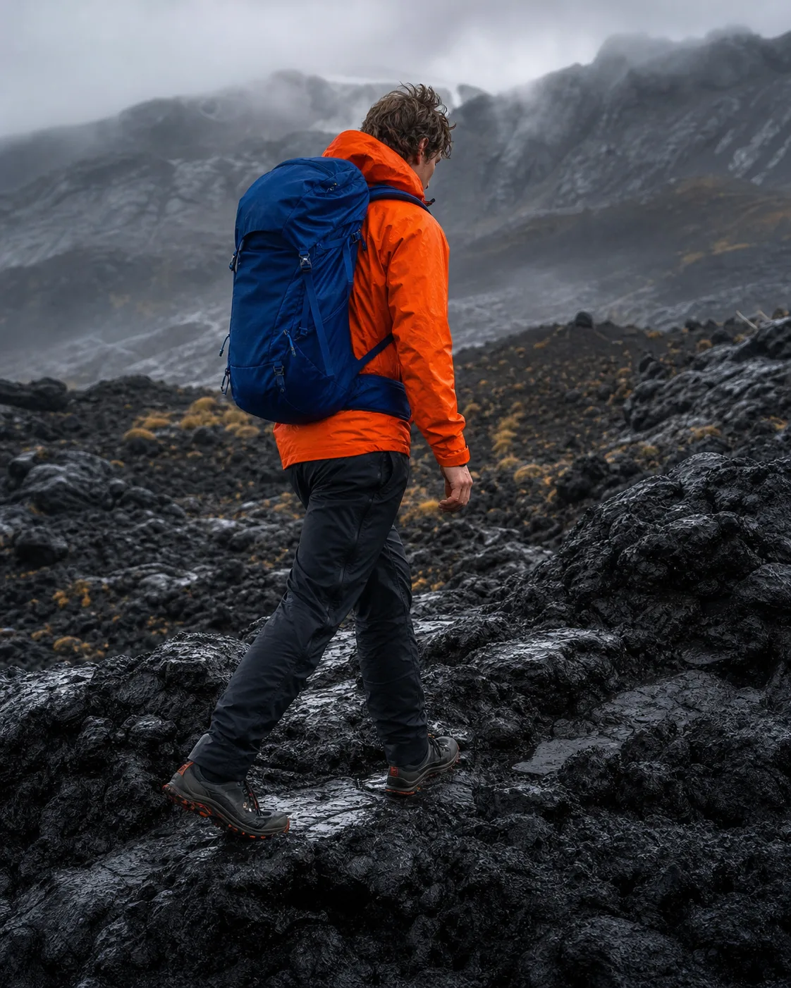 Hiker in an orange shell crossing wet volcanic terrain